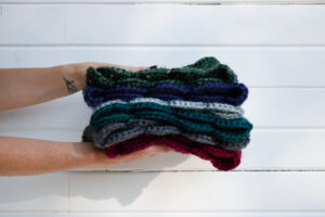 Two hands coming from the left of the photo are sandwiching a stack of hand knit cowls in multiple colors.