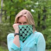 A blond woman is wearing a mint green crochet cowl and has it pulled up over her nose with her right hand.