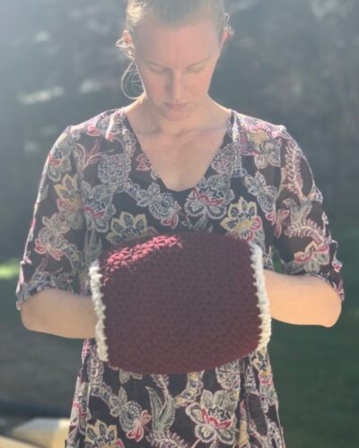 Liz, a blond woman, is wearing a floral dress and looking down at the burgundy hand warmer on her hands.