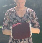 Liz, a blond woman, is wearing a floral dress and looking down at the burgundy hand warmer on her hands.