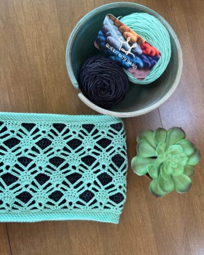 Mint green crochet cowl lying on a wood background. A bowl with yarn and a Queen City Yarn is next to the cowl.