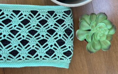 Mint green crochet cowl lying on a wood background. A bowl with yarn and a Queen City Yarn is next to the cowl.