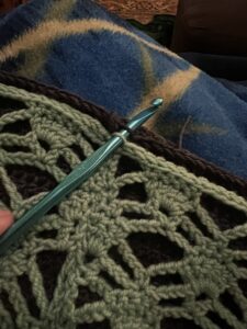 A crochet project with mint green and charcoal yarn. Crochet hook shown going through both layers of fabric.